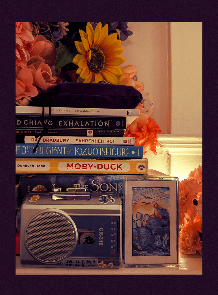 A cassette tape next to a small cassette player. Books like "Exhalation," by Ted Chiang; "Fahrenheit 451," by Ray Bradbury; and "Moby-Duck," by Donovan Hohn, are stacked behind.